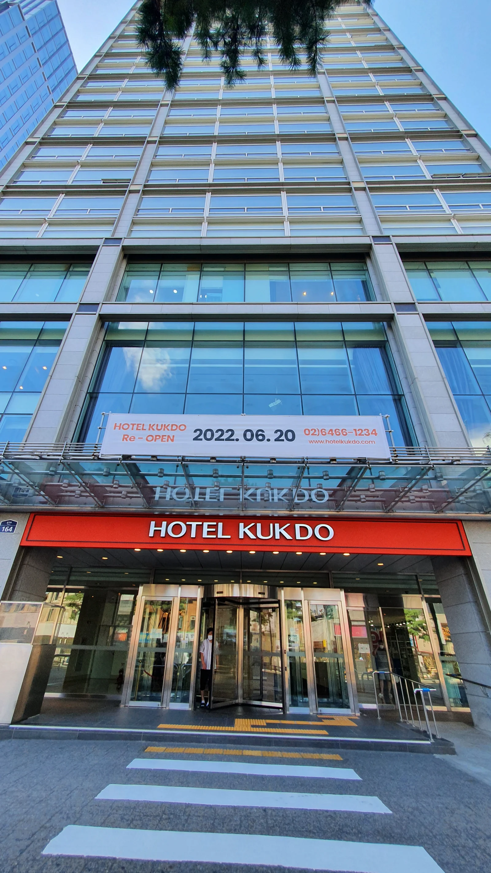 빨간 HOTEL KUKDO 간판과 회전문이 있는 호텔 정문, 그 위 유리창에 걸린 재개장 날짜 현수막
